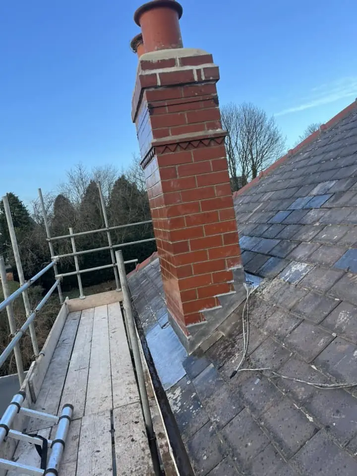 Tall brick chimney stack seen from the scaffold walkway, grey roof tiles beside it
